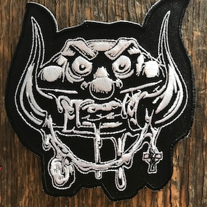 May include: Black and white embroidered patch of a cartoon monster with horns, dripping with a white liquid. The monster has a cross on its chest.