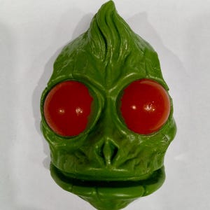 May include: A green, alien-like mask with large, red, round eyes. The mask has a textured surface and a pointed top. The overall design suggests a creature or character from science fiction or fantasy.