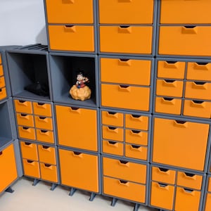 May include: A wall unit with multiple small drawers in a grid pattern. The drawers are orange and the unit is grey. A small figurine is on a shelf in the centre of the unit.