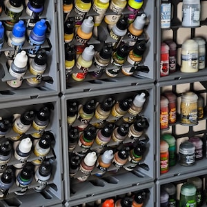 May include: A selection of miniature paint bottles and jars stored in grey containers. The bottles display a range of colours, such as blue, yellow, and brown. The jars are labelled with text. This image is ideal for miniature painting enthusiasts.