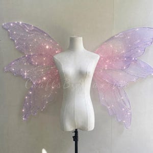 Op de afbeelding: Een paar grote, iriserende fee&euml;nvleugels in tinten lichtpaars en roze, met een glinsterende, doorschijnende textuur en ge&iuml;ntegreerde kleine witte lichtjes. De vleugels worden getoond op een eenvoudige witte torso-etalagepop.