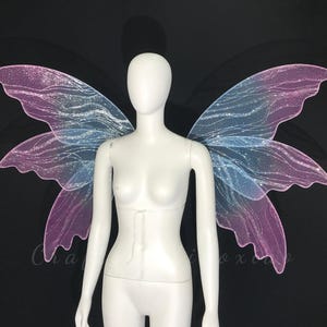 Op de afbeelding: Een witte etalagepop-torso met grote, iriserende fee&euml;nvleugels. De vleugels hebben een verloop van zachtroze en lichtblauw, geaccentueerd met een glinsterende glitterafwerking en ingewikkelde, vloeiende aderpatronen op het doorschijnende materiaal.