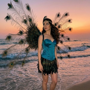 May include: A teal satin corset top paired with a short skirt featuring dark feathers. Large, expansive peacock feather wings are attached to the back. A thin, metallic headband with a small feather accent is worn across the forehead.