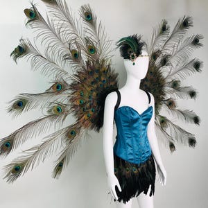 May include: A costume set featuring large, expansive wings constructed from iridescent peacock feathers, paired with a teal satin corset and a black feathered skirt. A matching peacock feather headpiece with a central green jewel is included.