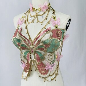 May include: A decorative butterfly-shaped top with pink, green, and gold embroidery. The top features a gold choker with pink butterfly accents and dangling gold chains. The butterfly design is detailed with intricate stitching and embellishments.
