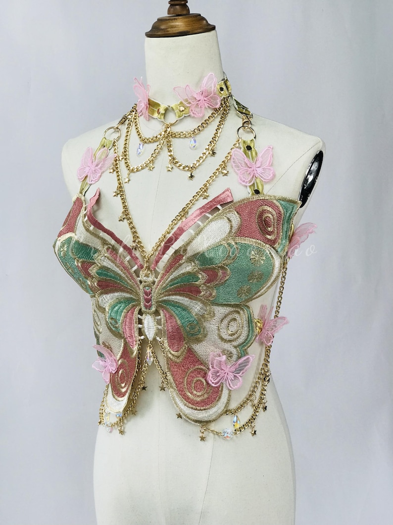 May include: A decorative butterfly-shaped top with pink, green, and gold embroidery. The top features a gold choker with pink butterfly accents and dangling gold chains. The butterfly design is detailed with intricate stitching and embellishments.