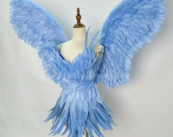 Puede incluir: Un par de alas de ángel azul claro hechas de plumas. Las alas están unidas a un maniquí y están extendidas. Las alas son un accesorio de disfraz popular para Halloween y otros eventos.