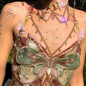 May include: A butterfly-themed top with gold chain accents. The top features an embroidered butterfly design in shades of green, pink, and gold. Additional details include a matching choker and decorative chains with star and gem pendants.