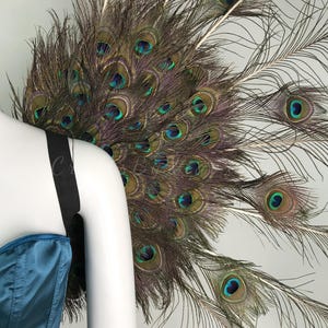 May include: A large fan-shaped accessory constructed from numerous iridescent peacock feathers featuring prominent blue and green eyespots. The feathers are arranged in a circular display and attached to a black elastic strap worn over a teal satin garment.