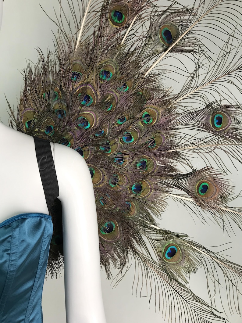 May include: A large fan-shaped accessory constructed from numerous iridescent peacock feathers featuring prominent blue and green eyespots. The feathers are arranged in a circular display and attached to a black elastic strap worn over a teal satin garment.