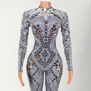 May include: A gray jumpsuit with long sleeves and a high neckline. The jumpsuit is embellished with mirrored, geometric shapes. The design features a pattern of hexagonal and rectangular mirrored pieces, creating a shimmering effect. The garment is form-fitting.