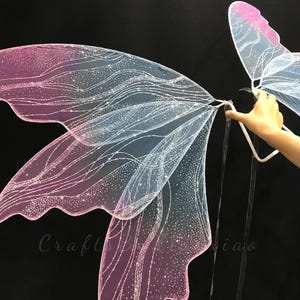 Op de afbeelding: Een paar iriserende elfenvleugels met een kleurverloop van roze en blauwe tinten. De vleugels zijn gemaakt van een doorschijnend materiaal met een delicaat, golvend zilveren glitterpatroon over het oppervlak en geschulpte randen.