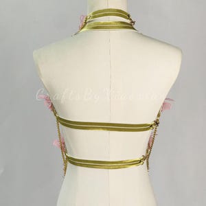 May include: A gold-colored body harness with three horizontal bands and a choker-style neckline. Delicate pink floral accents are placed on the harness. The harness is displayed on a mannequin against a neutral background. Gold chains connect the bands.