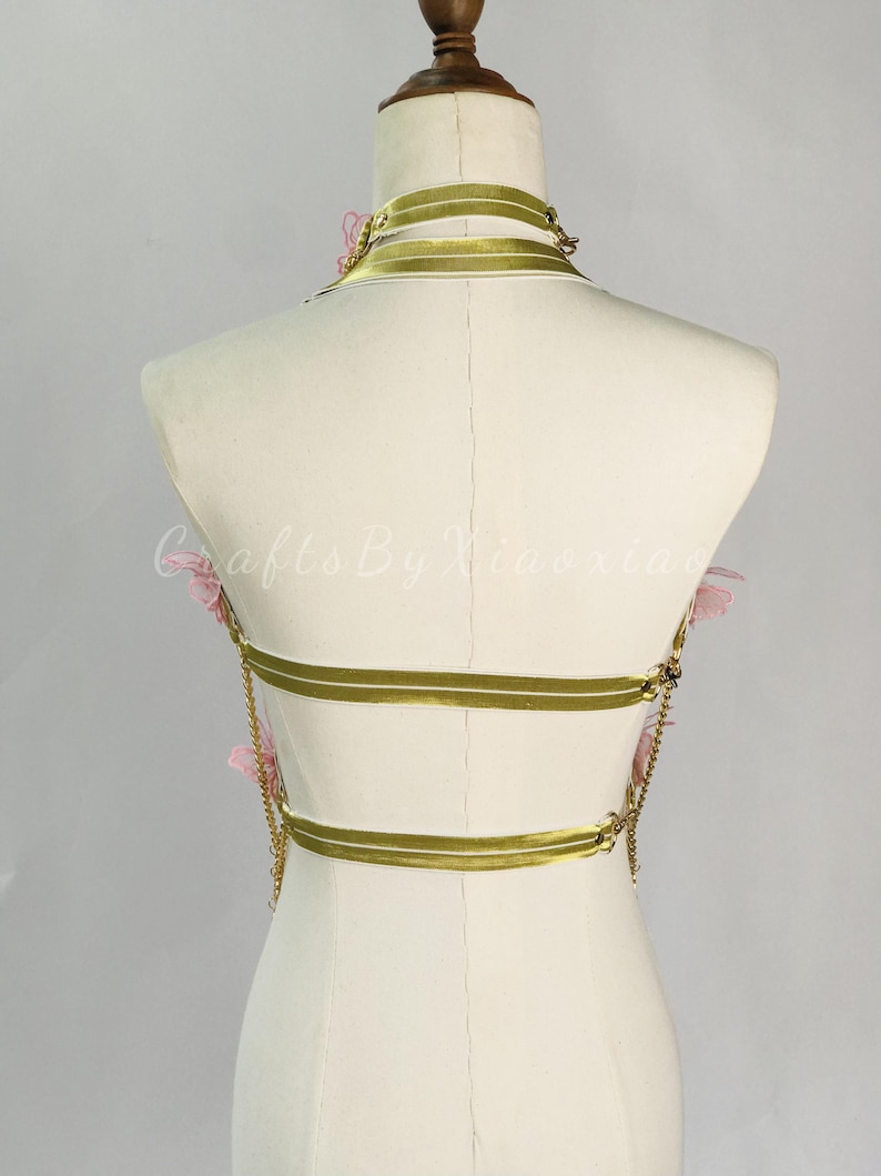 May include: A gold-colored body harness with three horizontal bands and a choker-style neckline. Delicate pink floral accents are placed on the harness. The harness is displayed on a mannequin against a neutral background. Gold chains connect the bands.