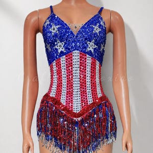 May include: A sequined bodysuit in a patriotic design, featuring a blue top with white stars and a red and white striped bodice. The bottom is trimmed with red and blue sequin fringe. The outfit is displayed on a mannequin.