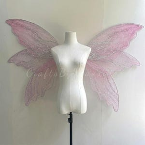 Op de afbeelding: Grote, doorschijnende lichtroze fee&euml;nvleugels met ingewikkelde zilveren glitteraders en geschulpte randen. De vleugels worden getoond op een witte paspop-torso en hebben een brede, uitgestrekte vorm met delicate, vloeiende rondingen en een glinsterende afwerking.
