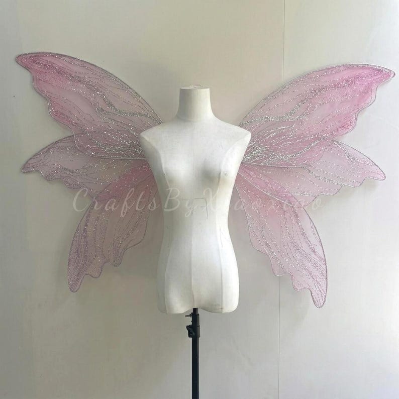 Op de afbeelding: Grote, doorschijnende lichtroze fee&euml;nvleugels met ingewikkelde zilveren glitteraders en geschulpte randen. De vleugels worden getoond op een witte paspop-torso en hebben een brede, uitgestrekte vorm met delicate, vloeiende rondingen en een glinsterende afwerking.