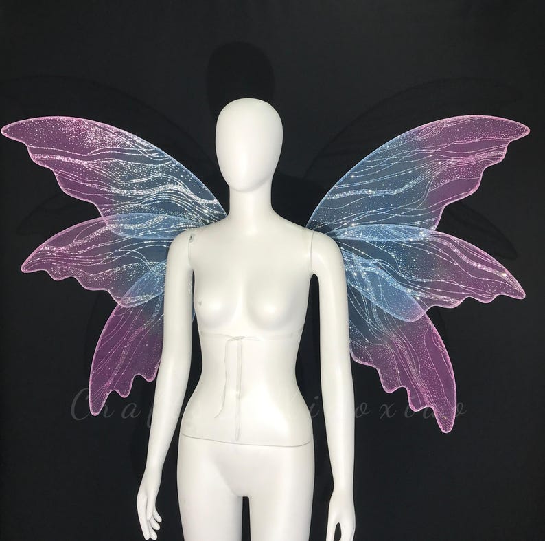 Op de afbeelding: Een witte etalagepop-torso met grote, iriserende fee&euml;nvleugels. De vleugels hebben een verloop van zachtroze en lichtblauw, geaccentueerd met een glinsterende glitterafwerking en ingewikkelde, vloeiende aderpatronen op het doorschijnende materiaal.