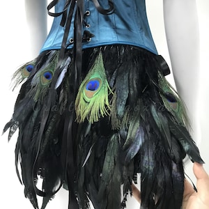 May include: A teal corset featuring a front black ribbon lace-up closure, paired with a skirt layered in black feathers and iridescent peacock eye feathers. The garment displays a structured bodice design with a dark, layered feather fringe.
