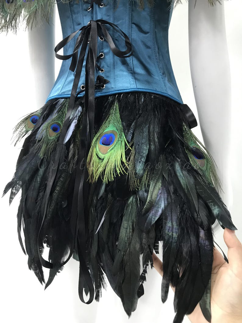 May include: A teal corset featuring a front black ribbon lace-up closure, paired with a skirt layered in black feathers and iridescent peacock eye feathers. The garment displays a structured bodice design with a dark, layered feather fringe.
