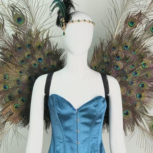 May include: A blue satin corset with a front hook closure, paired with large, expansive peacock feather wings. A decorative headpiece features a gold-toned band with green accents, a dark feather plume, and a dangling green teardrop-shaped crystal.