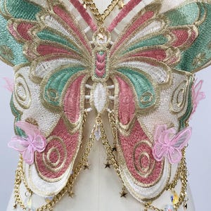 May include: Embroidered butterfly-shaped garment with pink, green, and gold detailing. Features gold chain accents with star and crystal charms, and delicate pink butterfly embellishments. The design is intricate and ornate.