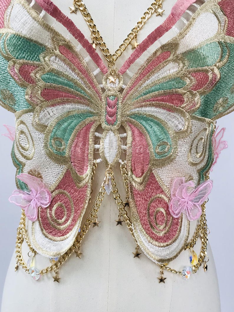 May include: Embroidered butterfly-shaped garment with pink, green, and gold detailing. Features gold chain accents with star and crystal charms, and delicate pink butterfly embellishments. The design is intricate and ornate.