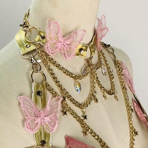May include: A gold-colored choker necklace with pink butterfly accents and dangling gold chains. The necklace features a gold band with pink butterfly appliqu&eacute;s and small star charms. The design includes a decorative bodice with pink and green embroidery.