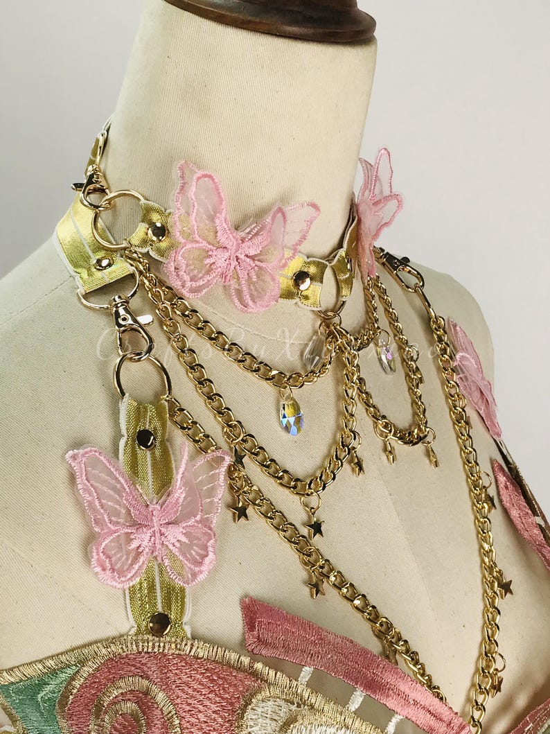 May include: A gold-colored choker necklace with pink butterfly accents and dangling gold chains. The necklace features a gold band with pink butterfly appliqu&eacute;s and small star charms. The design includes a decorative bodice with pink and green embroidery.