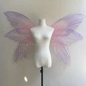 Op de afbeelding: Grote, iriserende fee&euml;nvleugels met een verloop van zachtroze en lichtpaarse tinten. De vleugels hebben een doorschijnende, glinsterende textuur met ingewikkelde aderpatronen en een fijne glitterafwerking, weergegeven op een witte torso-etalagepop.