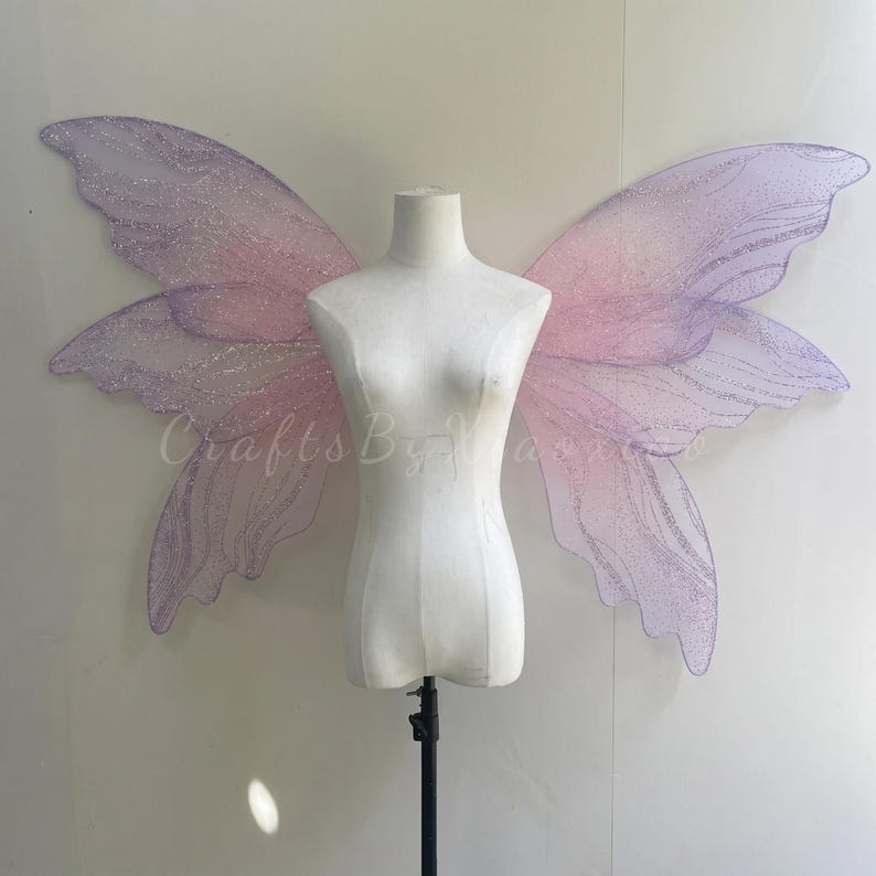 Op de afbeelding: Grote, iriserende fee&euml;nvleugels met een verloop van zachtroze en lichtpaarse tinten. De vleugels hebben een doorschijnende, glinsterende textuur met ingewikkelde aderpatronen en een fijne glitterafwerking, weergegeven op een witte torso-etalagepop.