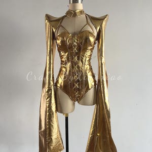 May include: A gold-colored bodysuit with long, flared sleeves and a high collar. The bodice features a corset-style design with lacing. The outfit is displayed on a mannequin. The text "Craft By Xiao" is visible.