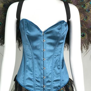 May include: A teal satin corset featuring a sweetheart neckline and a front metal hook-and-eye closure. The garment is accented with black elastic shoulder straps and a decorative trim of iridescent peacock feathers at the top and bottom edges.