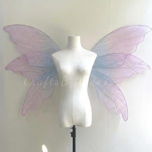 Op de afbeelding: Een paar grote, iriserende fee&euml;nvleugels met een verloop van zachte pastelroze en lichtblauwe tinten. De vleugels hebben een doorschijnende, glinsterende textuur met delicate aderachtige patronen en worden getoond op een witte etalagepop.