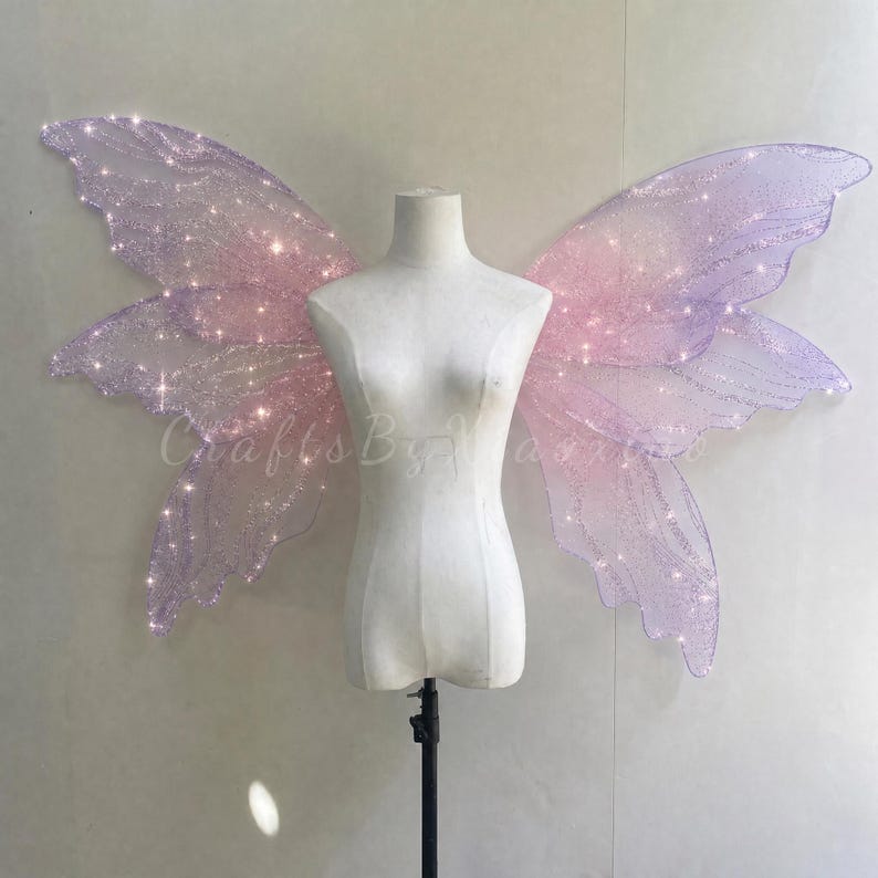Op de afbeelding: Een paar grote, iriserende fee&euml;nvleugels in tinten lichtpaars en roze, met een glinsterende, doorschijnende textuur en ge&iuml;ntegreerde kleine witte lichtjes. De vleugels worden getoond op een eenvoudige witte torso-etalagepop.