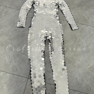 May include: A silver jumpsuit covered in small, reflective, hexagonal pieces. The long-sleeved garment has a high neckline and a fitted silhouette. The jumpsuit is designed to be worn as a costume or for performance.