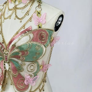 May include: A decorative butterfly-shaped chest harness with a matching choker. The harness features embroidered wings in pink, green, and gold, adorned with delicate pink butterfly accents and gold chains. The choker and harness are connected by gold chains.