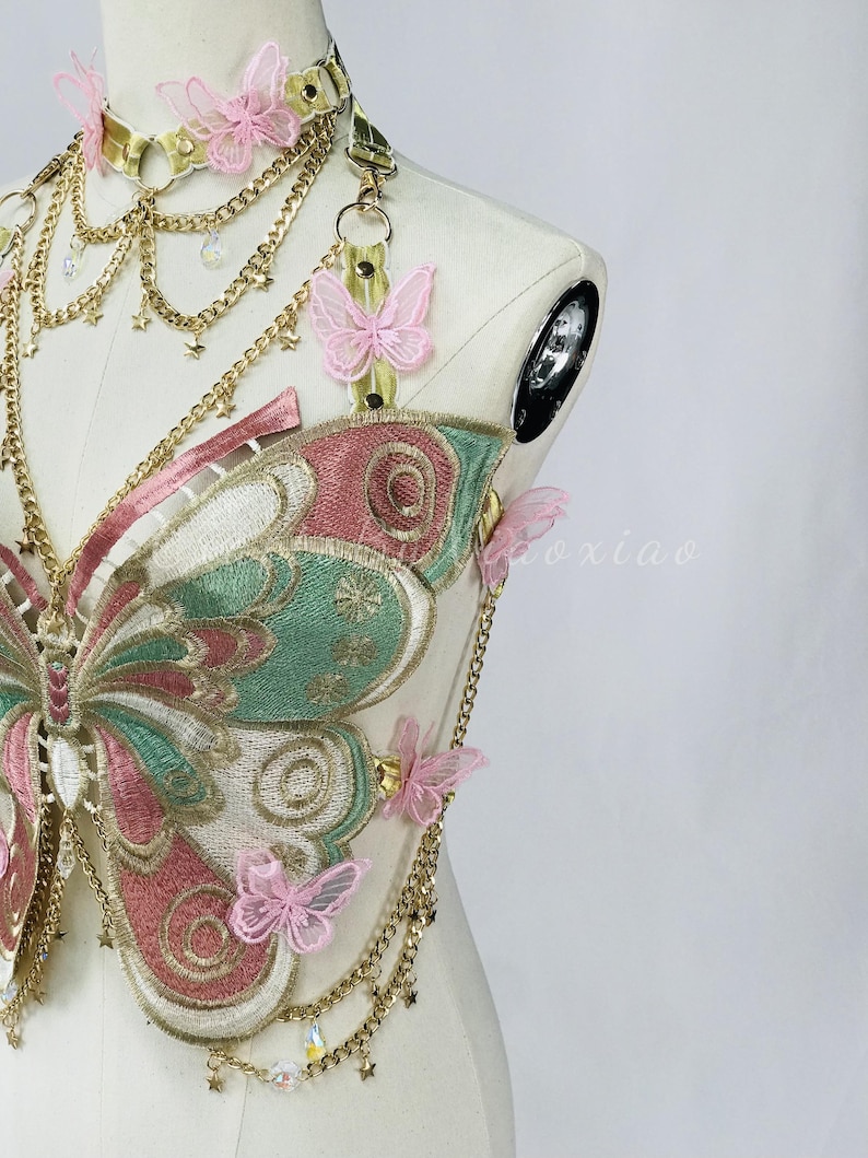 May include: A decorative butterfly-shaped chest harness with a matching choker. The harness features embroidered wings in pink, green, and gold, adorned with delicate pink butterfly accents and gold chains. The choker and harness are connected by gold chains.