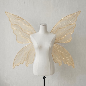 May include: A pair of large, translucent fairy wings with a gold glitter vein pattern and integrated small LED lights, displayed on a white mannequin torso. The wings feature a delicate, organic shape with scalloped edges and a shimmering finish.