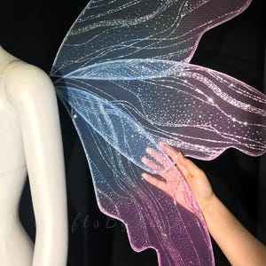 Op de afbeelding: Doorschijnende elvenvleugels met een kleurverloop van blauw naar roze. De vleugels zijn gemaakt van een fijn gaasmateriaal met een glinsterende, glitterende afwerking en ingewikkelde, golvende aderpatronen over het gehele oppervlak.