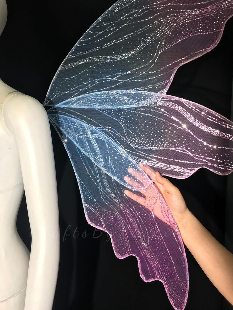 Op de afbeelding: Doorschijnende elvenvleugels met een kleurverloop van blauw naar roze. De vleugels zijn gemaakt van een fijn gaasmateriaal met een glinsterende, glitterende afwerking en ingewikkelde, golvende aderpatronen over het gehele oppervlak.