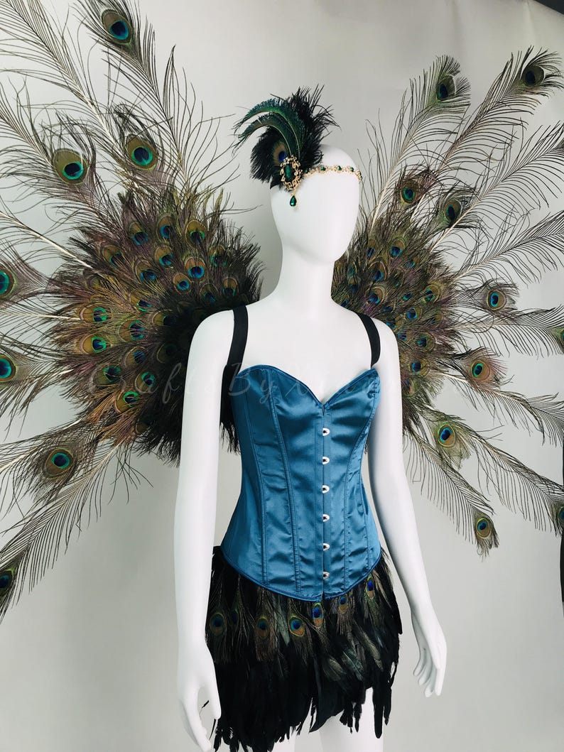 May include: A teal satin corset with a front hook closure, paired with a black feather skirt and large peacock feather wings. A matching headpiece features a green gemstone, black feathers, and a gold-toned chain across the forehead.