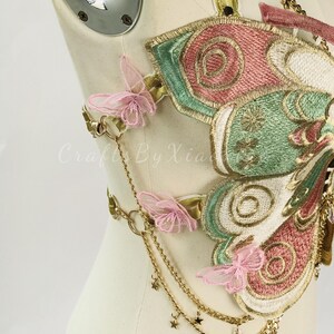 May include: Embroidered butterfly-shaped bodice with pink, green, and gold accents. Features delicate pink butterfly appliqu&eacute;s, gold chains, and star charms. The bodice is displayed on a mannequin.