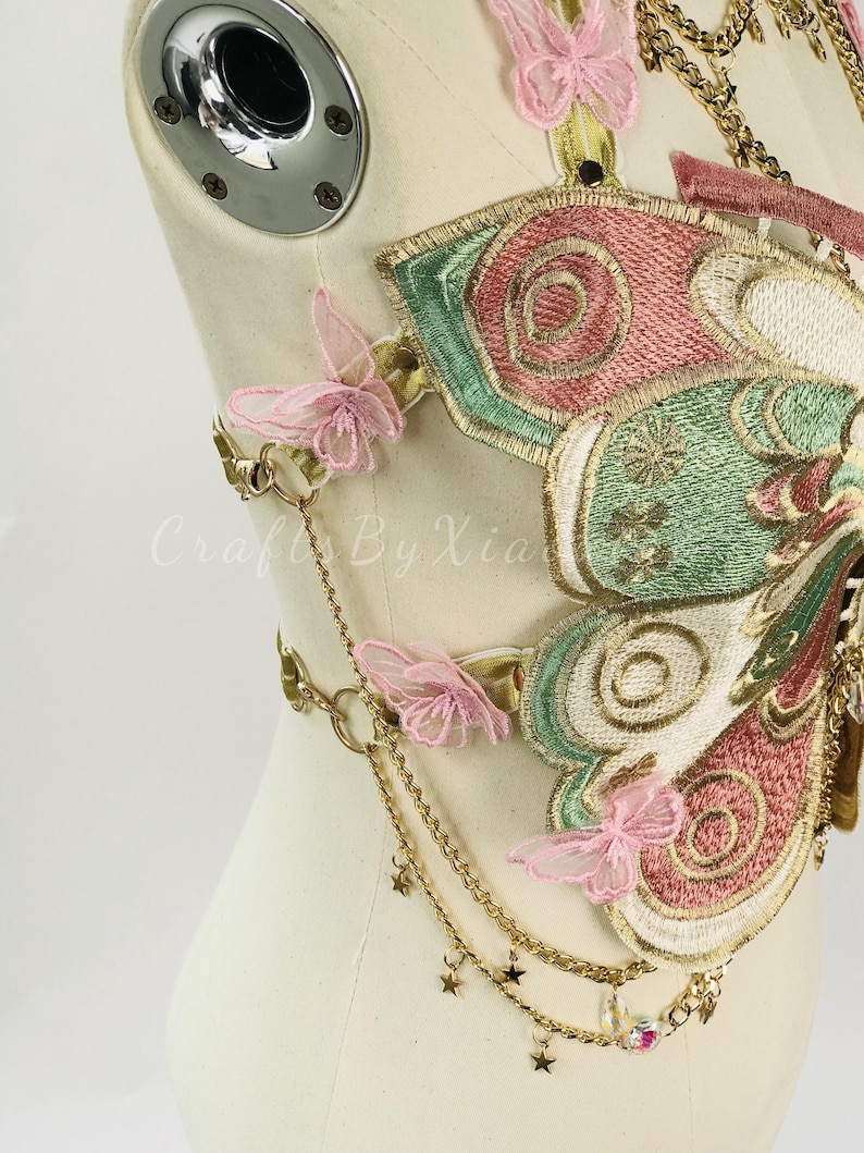 May include: Embroidered butterfly-shaped bodice with pink, green, and gold accents. Features delicate pink butterfly appliqu&eacute;s, gold chains, and star charms. The bodice is displayed on a mannequin.