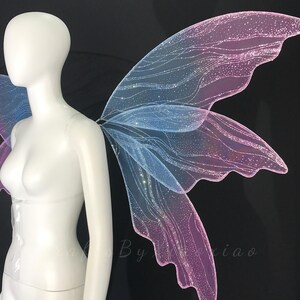 Op de afbeelding: Een paar iriserende elfenvleugels met een verloop van blauwe en roze tinten. De vleugels zijn gemaakt van een doorschijnend, glinsterend materiaal met een delicaat, aderachtig patroon en een fijne glitterafwerking op een etalagepop.