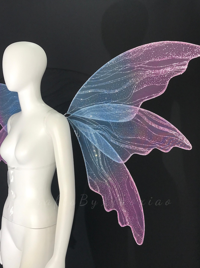 Op de afbeelding: Een paar iriserende elfenvleugels met een verloop van blauwe en roze tinten. De vleugels zijn gemaakt van een doorschijnend, glinsterend materiaal met een delicaat, aderachtig patroon en een fijne glitterafwerking op een etalagepop.