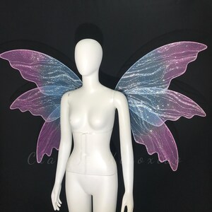 Op de afbeelding: Een paar grote, doorschijnende fee&euml;nvleugels met een verloop van zachtroze en lichtblauw. De vleugels hebben een delicaat, aderachtig patroon en zijn bedekt met een fijne, glinsterende glitter die het licht vangt tegen een zwarte achtergrond.