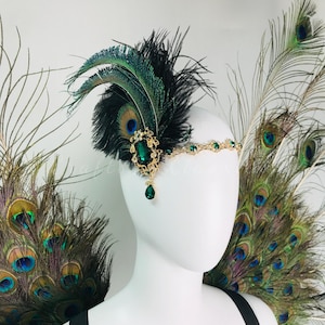 May include: A headpiece featuring a gold-toned metal band adorned with small green crystals. A large oval green crystal set in an ornate gold frame is attached to the side, accented with black feathers and iridescent peacock feathers.