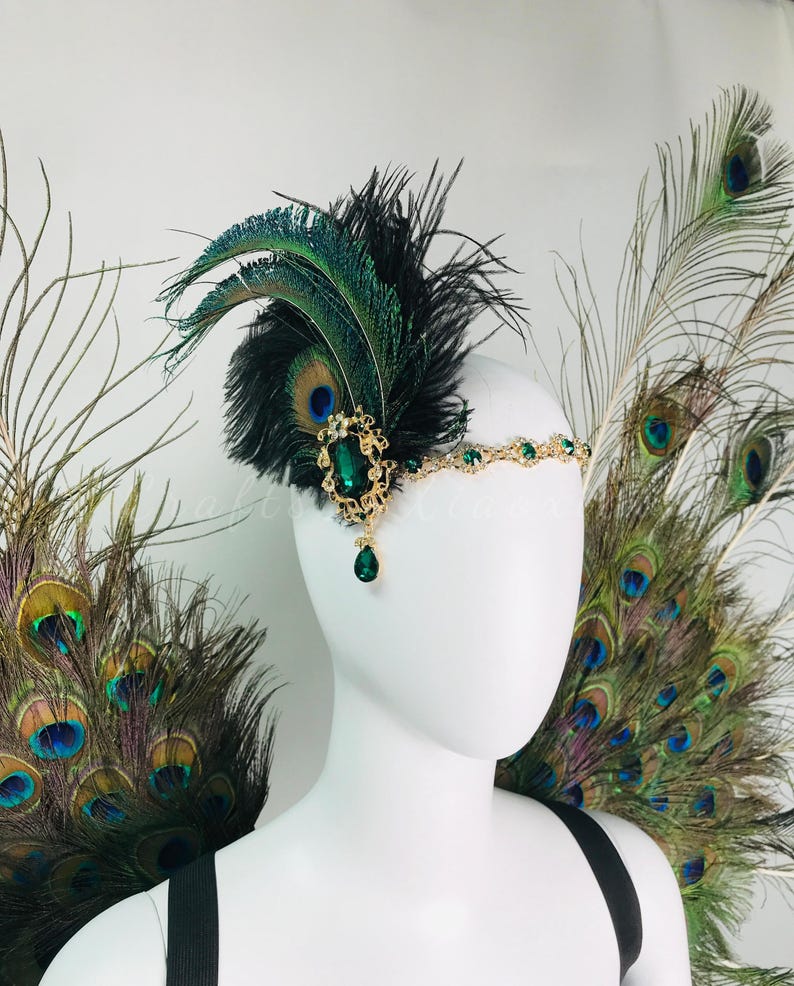 May include: A headpiece featuring a gold-toned metal band adorned with small green crystals. A large oval green crystal set in an ornate gold frame is attached to the side, accented with black feathers and iridescent peacock feathers.