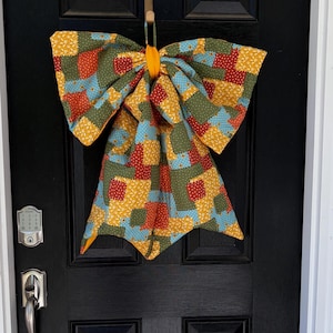 May include: A large fabric bow decoration featuring a patchwork pattern of quilted squares in shades of olive green, mustard yellow, light blue, and red with small floral and polka dot prints, centered with a solid mustard yellow fabric knot.